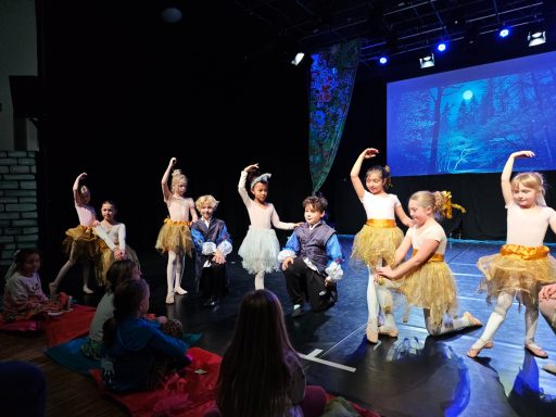 Tanzende Kinder in Kostümen mit Tüllröcken auf einer Bühne vor einem Projektor.