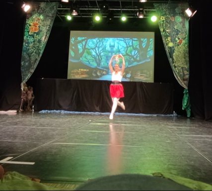Tänzerin in rotem Rock führt eine Aufführung auf einer Bühne vor einem Projektorbild auf.