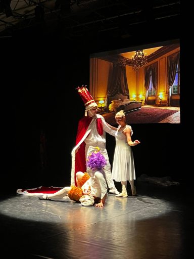 Ein königlicher Darsteller in buntem Outfit, umgeben von zwei weiteren Schauspielern.