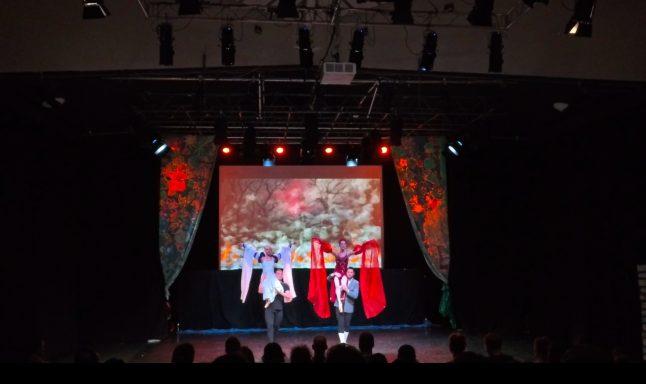 Zwei Performer auf der Bühne, buntes Licht und große Leinwand im Hintergrund.