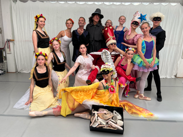 2025 "Die zertanzten Schuhe" Gruppenszene von Tänzern in bunten Kostümen, einige mit Requisiten, vor neutralem Hintergrund.