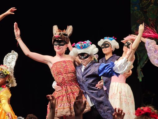 Mehrere Personen in festlicher Kleidung und Masken feiern auf einer Bühne.