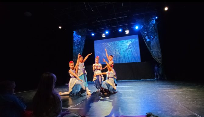 Tanzaufführung mit fünf Tänzern auf einer Bühne, beleuchtet von bunten Lichtern.