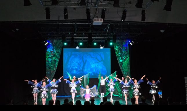 Tanzgruppe in bunten Kostümen auf einer Bühne mit Lichtern und Projektionen.