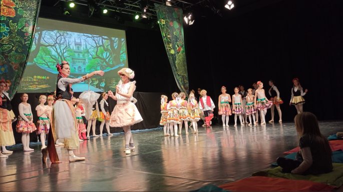 Bühne mit Tänzern in bunten Kostümen, einer führt eine Darbietung auf.